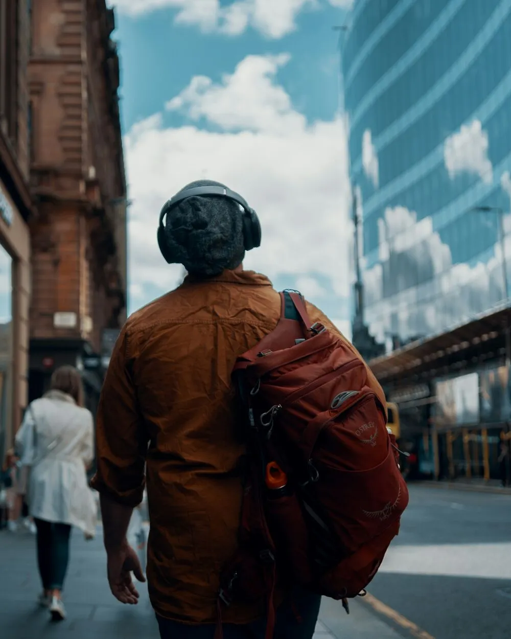 A man in headphones and an orange jacket walks down a city street between glass buildings